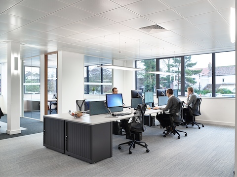 Three men seated in black Sayl office chairs work in a bright benching environment supported by Flo Monitor Arms.