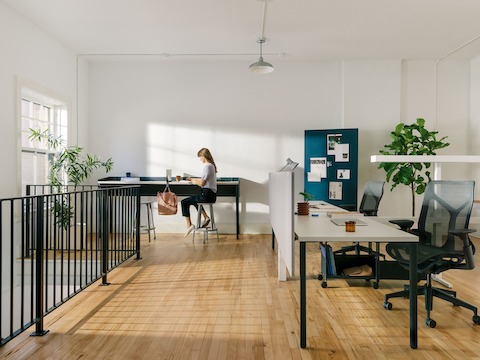 White OE1 Rectangular Tables in a workstations configuration, with a woman sitting at a blue OE1 Communal Table, and blue OE1 Mobile Easel and project board in the background.