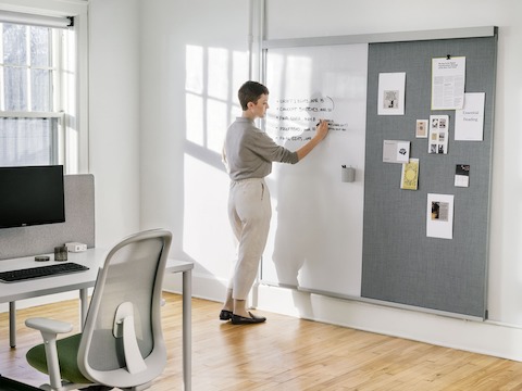 A woman writing on the OE1 Wall Rail and Board with an OE1 Rectangular Table and Lino Chair in the foreground.