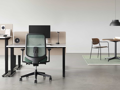 A black Verus Chair with dark green suspension back pulled up to a black Nevi Sit-Stand Desk with a Civic Table and Portrait Chair in the background.