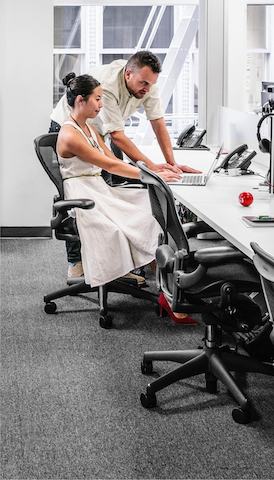A man and woman view a laptop computer in a benching setup featuring black Aeron office chairs.