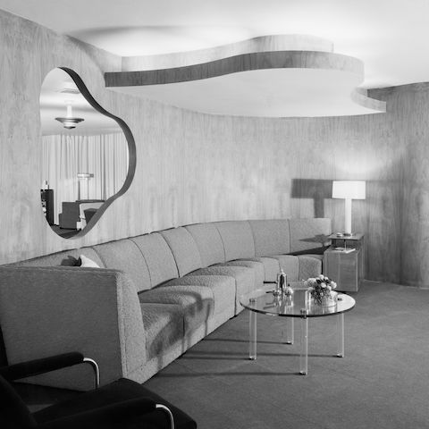 An archival black and white photo of Herman Miller's Chicago showroom in 1939, with curved wood walls with a freeform curvy cutout, upholstered sofa, and lucite coffee table and end table.