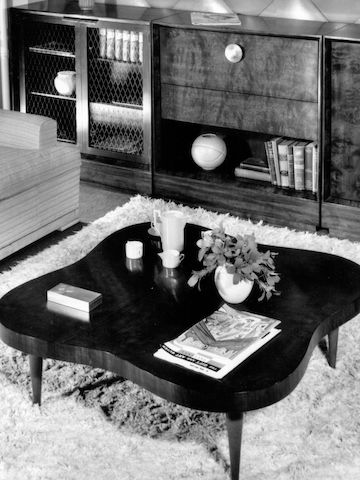 An archival black and white photo of the Paldao Table, a curvy organic-shaped low coffee table with various decor atop a shaggy rug, with wood cabinet and sofa nearby.