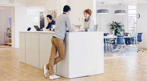 A man leans on a standing-height surface while speaking to a woman on a Setu Stool.